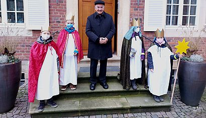 Bischof Genn steht mit vier Kindern, die als Sternsinger verkleidet sind, vor einem Gebäude.