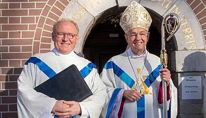 Ein Priester und ein Bischof stehen nebeneinander vor einem Eingang.