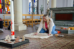 Eine Frau kniet auf dem Boden, vor ihr ist ein Tuch mit Sand ausgebreitet.