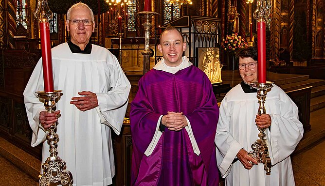 Zwei erwachsene Messdiener stehen mit Kerzenleuchtern in einer Kirche, zwischen ihnen ein Priester.
