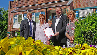 Bürgermeister Heiko Schmidt erhielt eine Ausgabe des Konzeptes von Gertrud Sivalingam, Pfarrer Günter Hoebertz und Natalie Heilen.  Sie stehen (von links) vor dem Rathaus von Sonsbeck, im Vordergrund blühen gelbe Blumen.