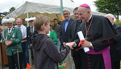 Bischof Felix Genn steht auf vor einem Zelt und reicht dem zehnjährigen Jonas die Hand, um ihm zum Geburtstag zu gratulieren. In der anderen Hand hält er ein zusammengerolltes Bild, das er zuvor geschenkt bekommen hat.