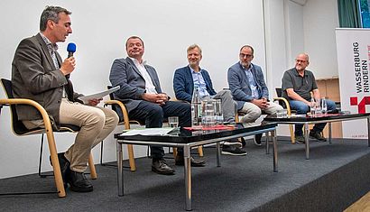 Fünf Männer sitzen auf einem Podium, einer von ihnen spricht in ein Mikrofon.