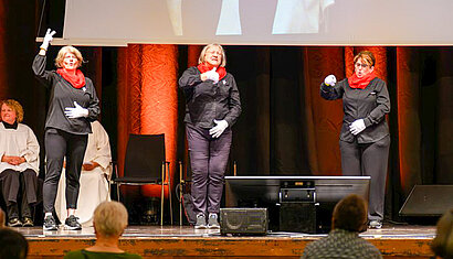 Auf einer Bühne stehen drei Frauen. Sie tragen schwarze Kleidung, rote Schals und weiße Handschuhe. Die Frauen haben ihre Hände erhoben, während sie lebhaft gebärden. 