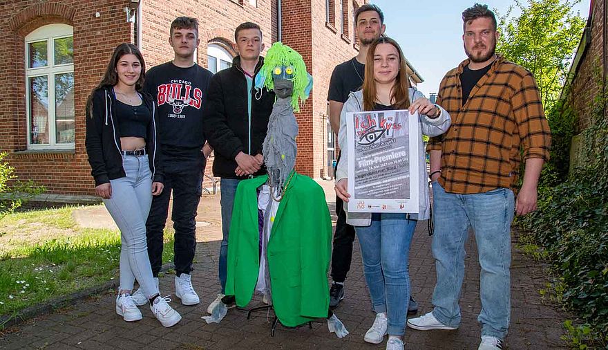 Sechs Menschen stehen um eine Kunstfigur mit grünen Haaren.