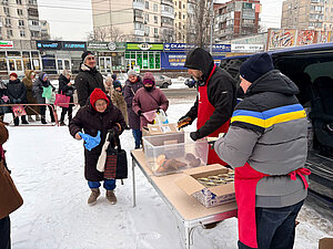 Auf dem Foto wird im Freien auf einer verschneiten Fläche eine Essensausgabe gezeigt. Mehrere Personen stehen in einer Schlange, während an einem Tisch Lebensmittel wie Brot und Konservendosen verteilt werden.