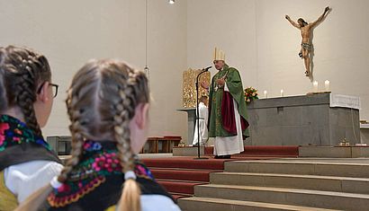 Bischof Genn steht an einem Mikrofon vor dem Altar und redet, während zwei junge Mädchen mit geflochtenen Haaren, die von hinten zu sehen sind, zuhören.