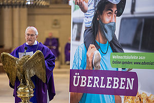 Ein Mann im violetten Messgewand steht an einem adlerförmigen Ambo neben einem Aktionsplakat der Adveniat-Weihnachtskation mit dem Titel "ÜberLeben in der Stadt".. 