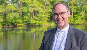 Weihbischof Rolf Lohmann steht an einem See. Im Hintergrund befinden sich grün belaubte Bäume.