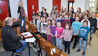 Zahlreiche Kinder stehen singend in einem Raum, vor ihnen sitzt Stephan Billen an einem Keyboard, im Hintergrund steht Petra Dobbelsteen auf einem Stuhl und lacht.