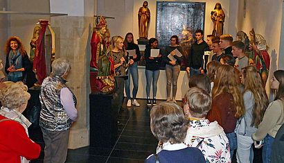Eine Gruppe aus Schülern und älteren Menschen steht im Stiftsmuseum, in dem Elisabeth Maas einen Vortrag hält.
