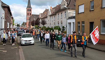 Ein langer Pilgerzug auf einer Straße, angeführt von einem Mann, der eine Caritas-Fahne trägt.