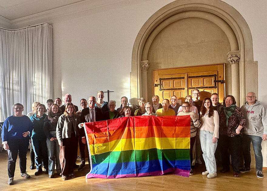 Eine Gruppe von rund 30 Personen hat sich zum Gruppenfoto hinter einer Regenbogenflagge versammelt.