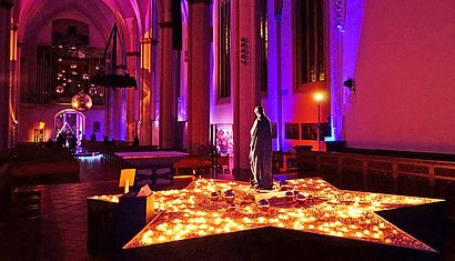 Der Innenraum der Überwasserkirche ist in buntes Licht getaucht, rund um eine Statue stehen brennende Opferkerzen.