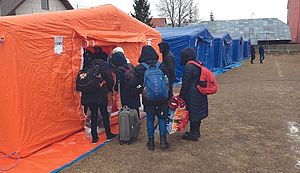 Fünf Männer und Frauen stehen bepackt mit Rucksäcken, Taschen und Kleinen Koffern am Eingang eines orangefarbenen Zeltes. Im Hintergrund befinden sich weitere Zelte.