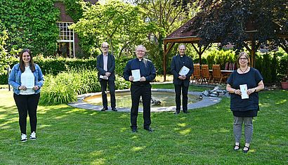 Zwei Frauen und drei Männer stehen mit Abstand auf einer schattigen Wiese und halten jeweils eine Broschüre in der Hand.