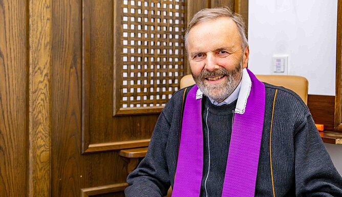 Ein Priester sitzt vor einer Holzwand mit einem hölzernen Geflecht.