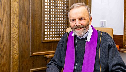 Ein Priester sitzt vor einer Holzwand mit einem hölzernen Geflecht.