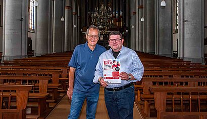 Zwei Männer stehen nebeneinander in einer Kirche, eine hat ein Plakat in der Hand.