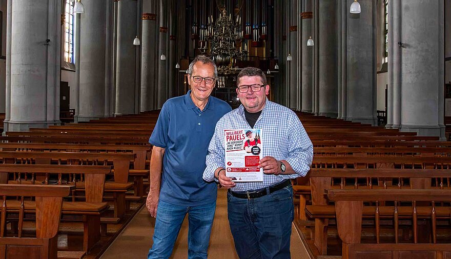 Zwei Männer stehen nebeneinander in einer Kirche, eine hat ein Plakat in der Hand. Zwei Männer stehen nebeneinander in einer Kirche, eine hat ein Plakat in der Hand.