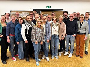 Eine Gruppe von 22 Erwachsenen steht dicht nebeneinander in einem hellen Innenraum und blickt in die Kamera. Die Personen wirken gemischt in Alter und Geschlecht, tragen überwiegend legere Kleidung wie Jeans, Pullover und Hemden und lächeln größtenteils. Einige tragen Brillen, eine Person hat einen auffälligen Bart. Im Hintergrund sind Stellwände mit angehefteten Notizen oder Plakaten zu sehen, was auf einen Workshop oder ein Seminar hindeutet. Über einer Tür hängt ein grünes Notausgangsschild. Der Boden besteht aus hellem Holz. Eine Gruppe von 22 Erwachsenen steht dicht nebeneinander in einem hellen Innenraum und blickt in die Kamera. Die Personen wirken gemischt in Alter und Geschlecht, tragen überwiegend legere Kleidung wie Jeans, Pullover und Hemden und lächeln größtenteils. Einige tragen Brillen, eine Person hat einen auffälligen Bart. Im Hintergrund sind Stellwände mit angehefteten Notizen oder Plakaten zu sehen, was auf einen Workshop oder ein Seminar hindeutet. Über einer Tür hängt ein grünes Notausgangsschild. Der Boden besteht aus hellem Holz.