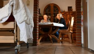 Zwei Frauen sitzen in einer Wohnung an einem Tisch und sprechen miteinander. Im Vordergrund ist das Fußteil eines Pflegebettes sichtbar.