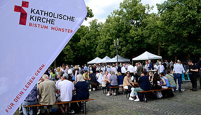 Auf dem Domplatz sitzen Menschen an Biertischen, im Hintergrund sind Pavillons aufgestellt. Im Vordergrund sieht man eine Werbefahne mit dem Logo des Bistums Münster.
