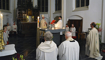 Weihbischof Rolf Lohmann steht hinter dem Altar, auf dem fünf Feuer brennen, im Vordergrund knien mehrere Personen.