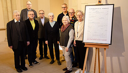 Die Mitglieder der Arbeitsgruppe stehen neben dme Plakat im Dom, auf dem oben zu lesen ist: "Im Anfang war die Tat - Missbrauch" und unten: "Eine offene Wunde im Dom zu Münster".