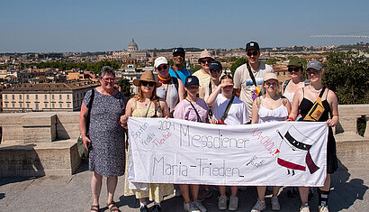Eine Messdiener-Gruppe steht mit einem selbst bemalten Tuch an einer Sehenswürdigkeit in Rom. Auf dem Tuch steht: Messdiener Maria-Frieden goes Rom.