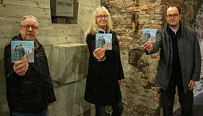 In der Krypta des Xantener Doms stehen an der Grubenlampe eine Frau und zwei Männer und halten ein Bilderbuch in die Kamera.