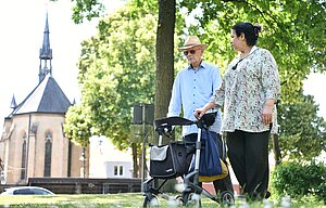 Ein älterer Mann mit Sonnenhut und Rollator spaziert gemeinsam mit einer Frau durch einen Park; im Hintergrund ist eine Kirche zu sehen.