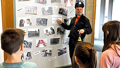 Schulseelsorger Markus van Berlo trägt eine Polizeimütze, in der linken Hand hält er eine Glocke. Er zeigt auf eine Wand mit Fotos, drei Kinder stehen mit dem Rücken zur Kamera und hören van Berlo zu.