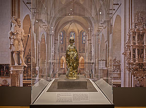 Die goldfarbene Statuette der heiligen Agnes, ausgestellt in einer Glasvitrine vor einem Bild, das eine Innenansicht des Langschiffs des St.-Paulus-Doms zeigt.