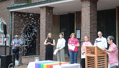 Die Vorbereitungsgruppe des Gottesdienstes steht hinter dem Altar, der mit einer Regenbogenfahne geschmückt ist. Im Hintergrund steigen Seifenblasen auf. 