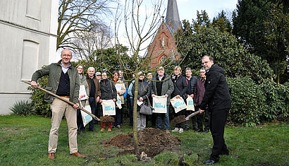 Zwei Männer schütten mit einer Schaufel Erde auf einen frisch eingepflanzten Baum, im Hintergrund stehen Gemeindemitglieder. Am Horizont ist eine Kirche zu sehen.