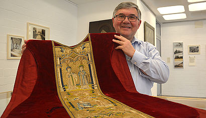 Pastor Alois van Doornick steht in einem Raum des Museums. Vor ihm ausgebreitet liegt ein Messgewand aus rotem Samt mit goldenen Stickereien, das er an der obren Hälfte anhebt.