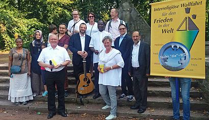 Zahlreiche Menschen stehen im Park neben einem gelben Plakat, auf dem "Interreligiöse Wallfahrt für den Frieden in der Welt" zu lesen ist.