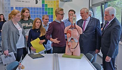 Mehrere Personen stehen in einem Raum, an dessen Wand ein Poster mit der Periodensystem hängt, und reden miteinander. Auf dem Tisch vor ihnen steht ein medizinisches Körpermodell.