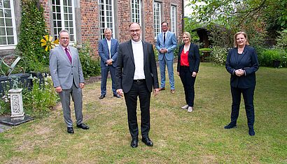 Zwei Frauen und vier Männer stehen mit Abstand zueinander in einem garten vor einem Backsteingebäude.