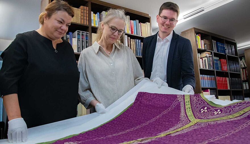 Zwei Frauen und eine Mann blicken auf ein auf einem Tisch liegendes Messgewand.