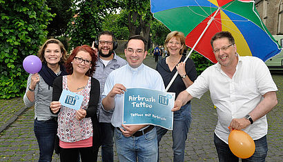 Das Organisationsteam, bestehend aus (von links) Maxi Decker, Maike Holtkamp, Martin Deckers, Kaplan Christoph Potowski, Simone Althoff und Daniel Frinken steht hinter dem Xantener Dom. Die Personen halten Gegenstände wie Luftballons, Hinweisschilder und einen bunten, aufgespannten Sonnenschirm in der Hand.