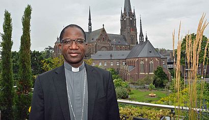 Erzbischof Laurent Lompo steht auf einem Balkon, hinter ihm ist die Marienbasilika Kevelaer zu sehen.