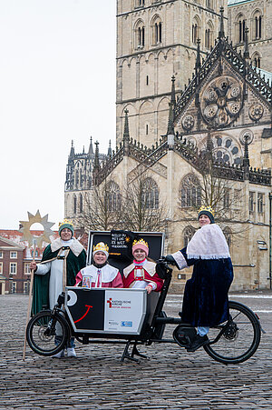 Vier Sternsingerinnen und Sternsinger in festlichen Gewändern und mit Kronen stehen mit einem Lastenrad auf dem Domplatz. Im Hintergrund erhebt sich der St.-Paulus-Dom in Münster. Eine Person hält einen großen Sternenstab, zwei Kinder sitzen in der Ladefläche des Fahrrads. Auf dem Lastenrad steht „Katholische Kirche Bistum Münster“. Vier Sternsingerinnen und Sternsinger in festlichen Gewändern und mit Kronen stehen mit einem Lastenrad auf dem Domplatz. Im Hintergrund erhebt sich der St.-Paulus-Dom in Münster. Eine Person hält einen großen Sternenstab, zwei Kinder sitzen in der Ladefläche des Fahrrads. Auf dem Lastenrad steht „Katholische Kirche Bistum Münster“.