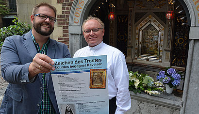 Dr. Bastian Rütten (links) und Wallfahrtsrektor Gregor Kauling stehen vor der Gnadenkapelle in Kevelaer. Rütten hält ein Plakat in der Hand, auf dem "Zeichen des Trostes" zu lesen ist.