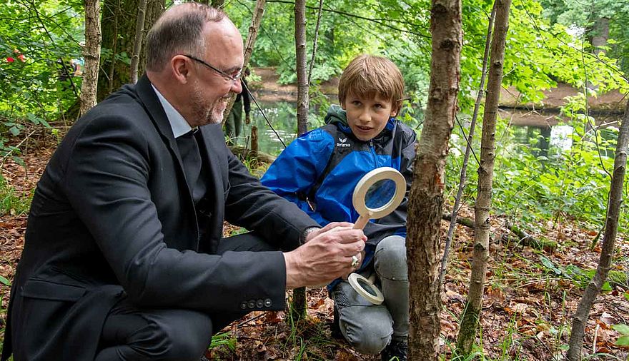 Ein Mann und ein Junge knien auf dem Waldboden, der Mann hat eine Lupe in der Hand.