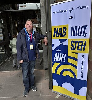 Ein Mann steht vor einem Kirchenportal, rechts neben ihm steht ein Katholikentagsbanner. Ein Mann steht vor einem Kirchenportal, rechts neben ihm steht ein Katholikentagsbanner.