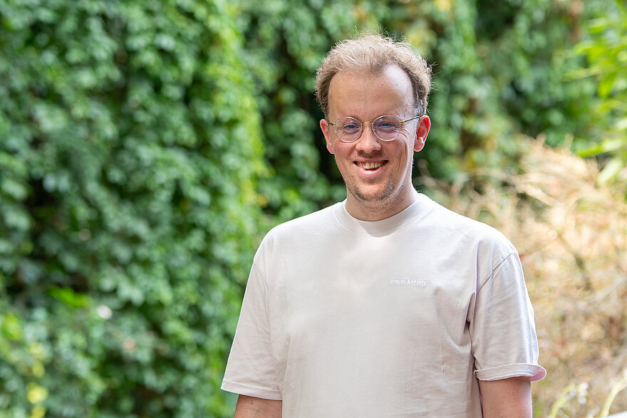 Jakob Rauschel schaut lächelnd in die Kamera und steht vor einer Hecke.