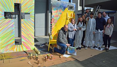 Vor zwei bunten Graffitibildern stehen mehrere Studentinnen und Studenten, einige halten Spraydosen in der Hand und tragen weiße Schutzanzüge. In der Mitte kniet der Künstler Mika Springwald.