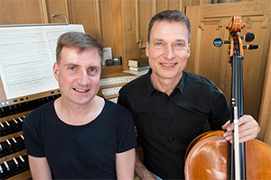 Lutz Wagner und Thomas Schmitz sitzen nebeneinander vor einer Orgel. Lutz Wagner hält ein Violoncello in der linken Hand.